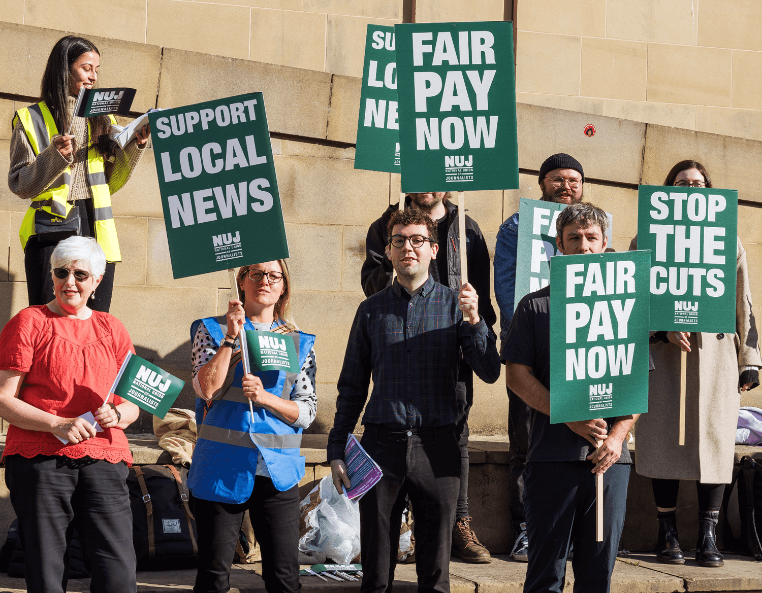 Journalists on a picket line holding greenplacards saying fair pay now, support local news