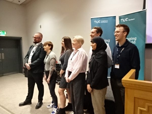A group of men and woman stand together, posing for a picture, in front of two blue stand-up banners for the TUC