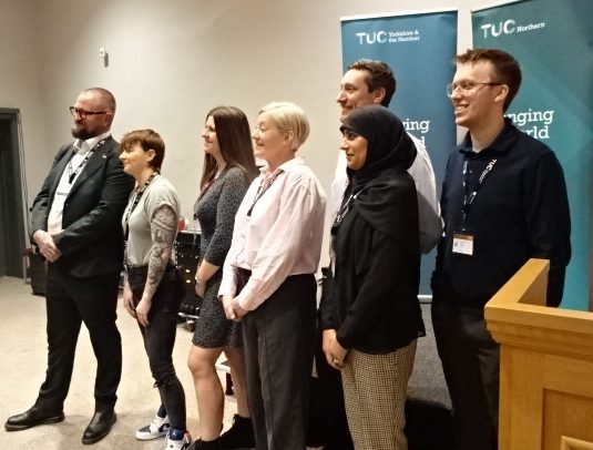 A group of men and woman stand together, posing for a picture, in front of two blue stand-up banners for the TUC