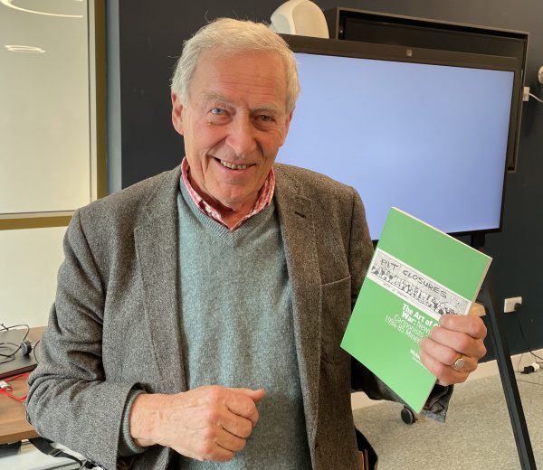 Smiling man with short white hair, teal jumper and tweed jacket holding a green book.