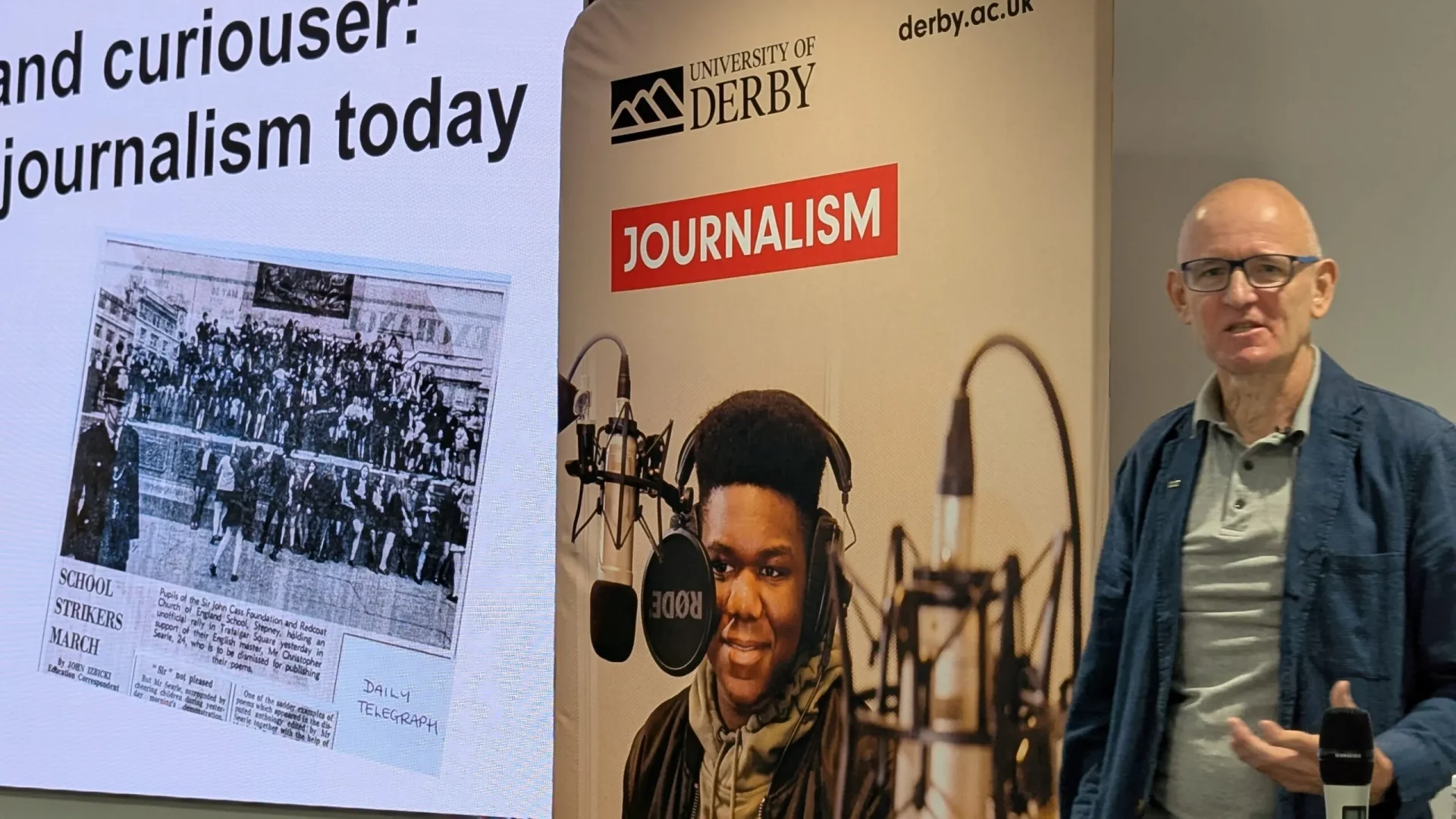 A man stands beside a University of Derby journalism display, speaking or presenting. Behind him is a large screen showing a newspaper clipping titled "SCHOOL STRIKERS MARCH" from the Daily Telegraph and a presentation slide that reads "and curiouser: journalism today". A promotional banner beside him features a young man speaking into a professional microphone in a radio studio.