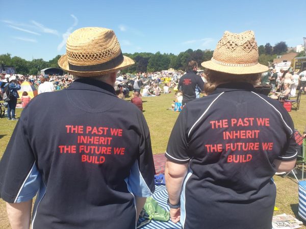 The backs of two people wearing straw hats and black t-shirts with red wording reading: "The past we in inherit, the future we build". They are in greenspace with crowds of people visible in the background