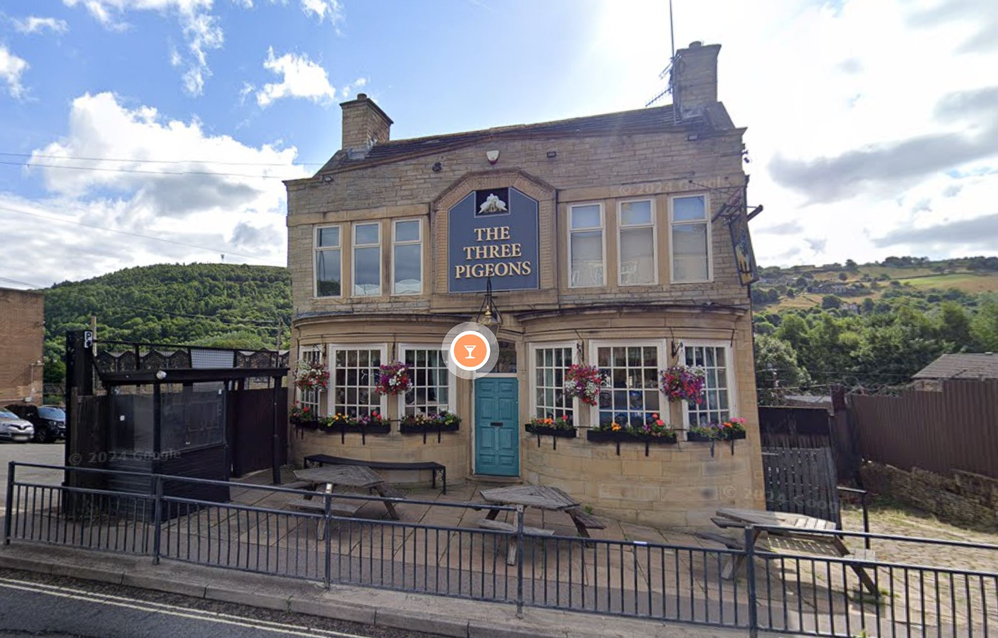 Exterior of a stone-built pub named “The Three Pigeons,” featuring large windows with flower boxes and hanging baskets of colorful flowers. Wooden benches and tables are arranged outside for seating. The pub is surrounded by green hills under a partly cloudy sky.
