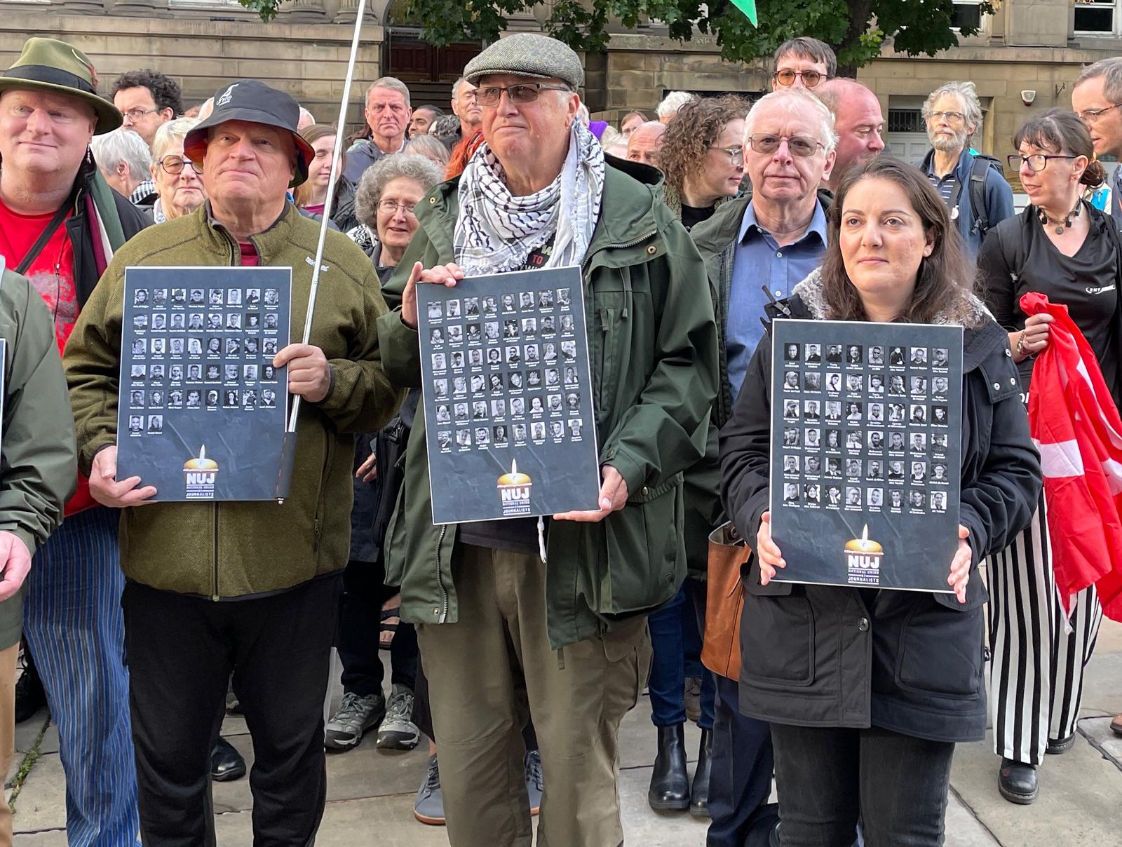 A group of people at a vigil, with three individuals at the front holding boards displaying black-and-white portraits of journalists. The boards have the NUJ (National Union of Journalists) logo at the bottom.
