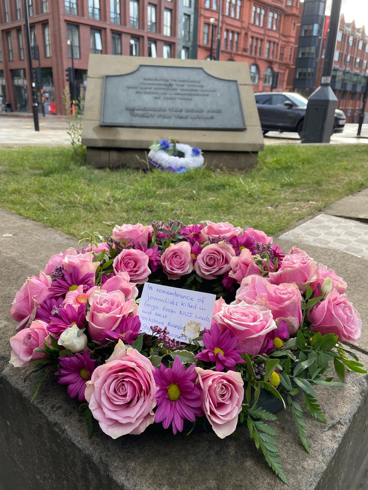 A floral wreath made of pink roses and purple daisies placed on a stone memorial. A handwritten note in the middle reads: “In remembrance of journalists killed in Gaza, from NUJ Leeds and NUJ Yorkshire Branch. Stop killing journalists.” A stone plaque in the background is engraved with a remembrance message.