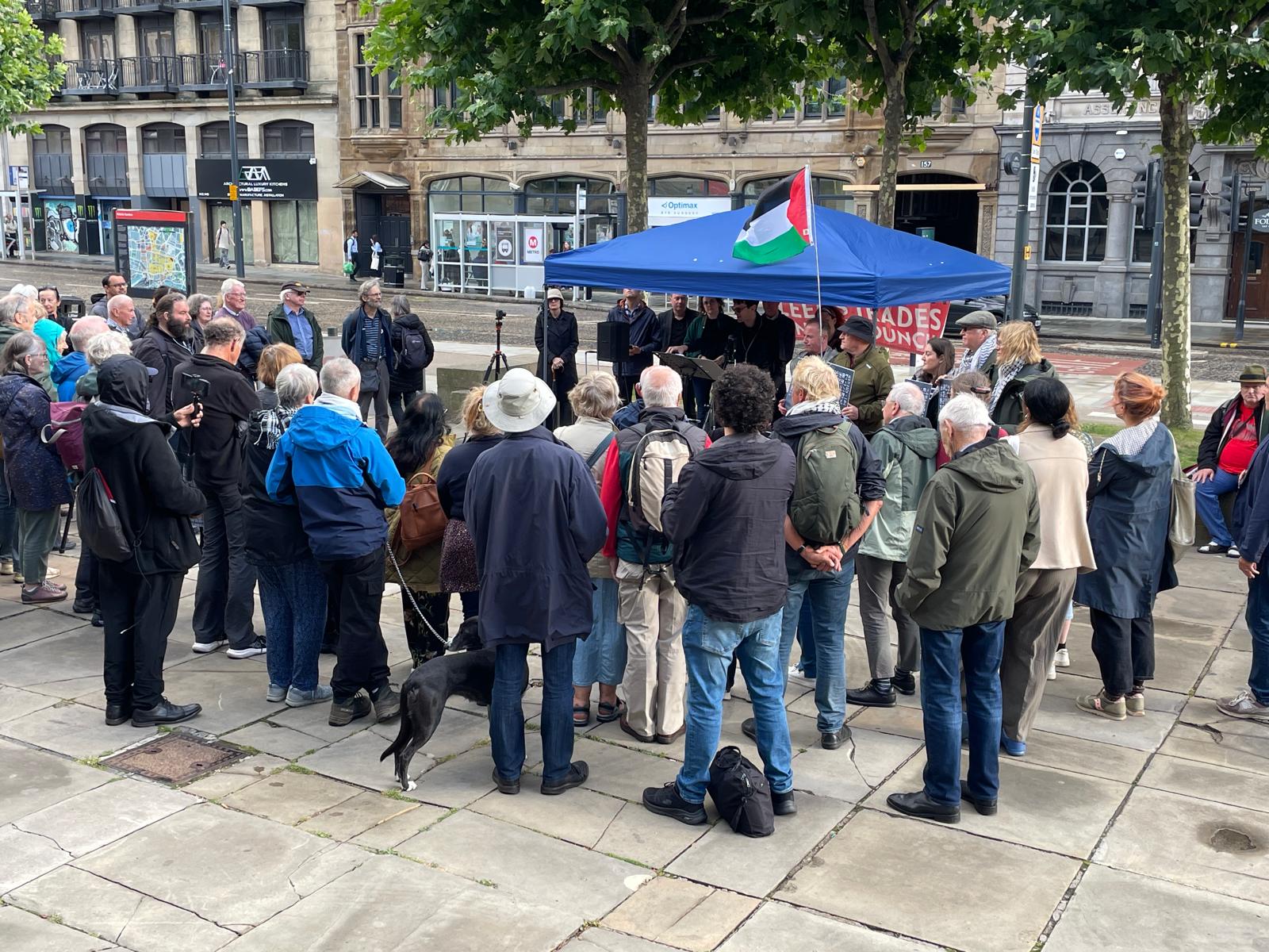 A crowd gathered outdoors around a small stage covered by a blue canopy with a Palestinian flag flying. The canopy displays a banner reading “Leeds Trades Council.” People are standing closely together, some with cameras, while a dog is also visible among the crowd.