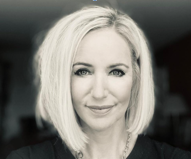 Black and white image of Leigh Purves looking into the camera and smiling. Her shoulder length hair is parted to the left. She is wearing a fark top and a chain necklace