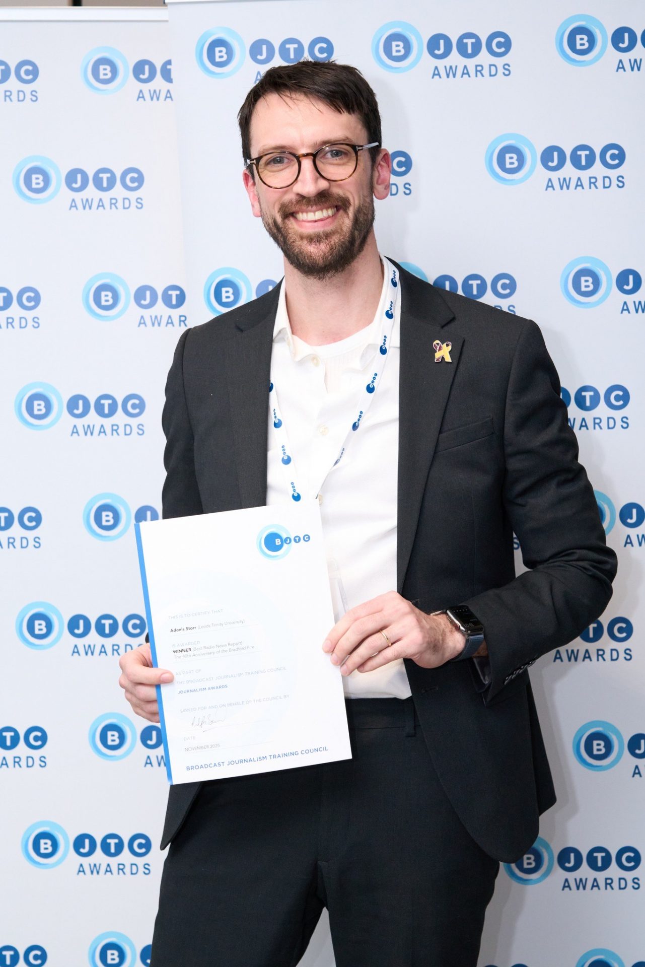 Adonis, wearing a white shirt and black suit jacket, smiles as he looks into the camera. He is standing in front of a background featuring the BCTJ logo
