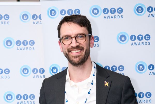 Adonis, wearing a white shirt and black suit jacket, smiles as he looks into the camera. He is standing in front of a background featuring the BCTJ logo