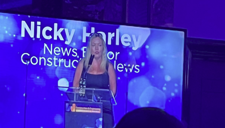 A woman stands at a podium speaking into a microphone at a formal event. Behind her, a large screen displays the name “Nicky Harley” and additional text. The stage lighting is purple and blue, creating a professional, celebratory atmosphere.