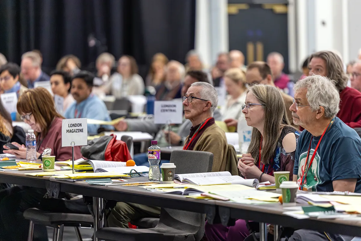 Members sitting at rows of tables looking towards an unseen speaker. Documents are spread out on the tables in front of them and names of different branches are displayed on various tables