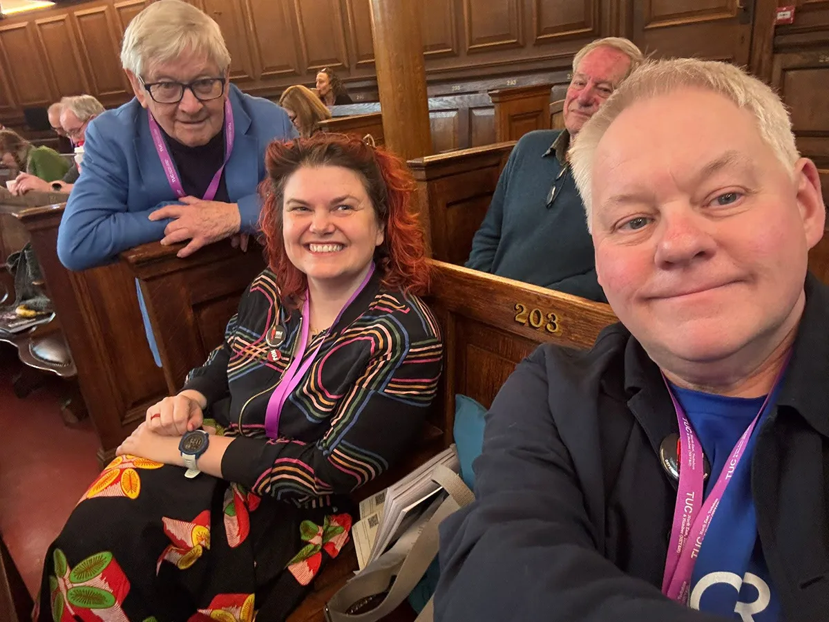 A selfie with Andy Walker in the foreground and Katie Hall sitting along from him on a wooden pew, while Barry White leans his crossed arms on the end of the pew and also looks towards the camera