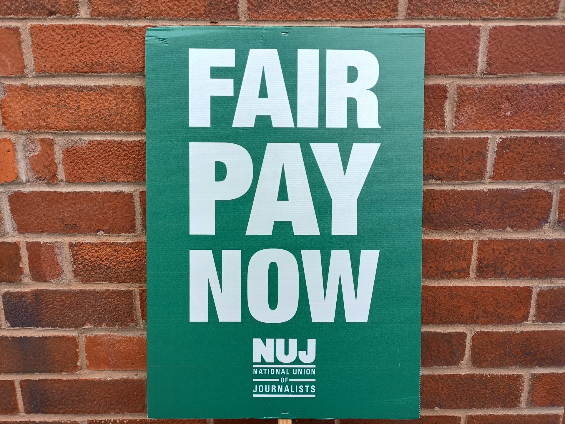 A green NUJ placard is propped against a brick wall. The white wording reads FAIR PAY NOW and the NUJ logo is below