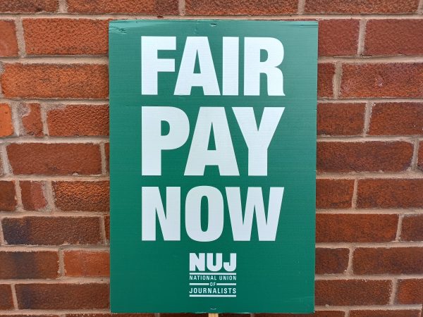 A green NUJ placard is propped against a brick wall. The white wording reads FAIR PAY NOW and the NUJ logo is below